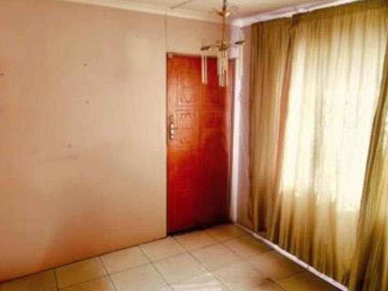 An empty living room featuring tiled flooring, a central ceiling light fixture, and a solid wood door, offering a blank canvas for renovation.