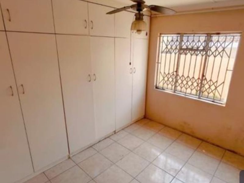 A vacant bedroom featuring extensive built-in cupboard storage, tiled flooring, a ceiling fan, and a window fitted with security bars.