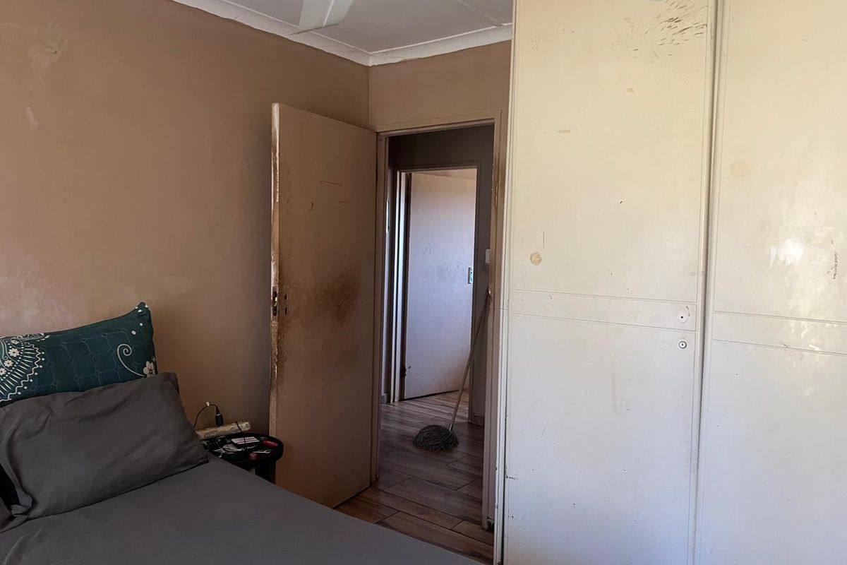 A bedroom featuring built-in cupboards and a ceiling fan, currently showing significant wear and tear on the walls and doors.