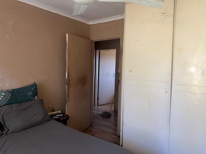 A bedroom featuring built-in cupboards and a ceiling fan, currently showing significant wear and tear on the walls and doors.