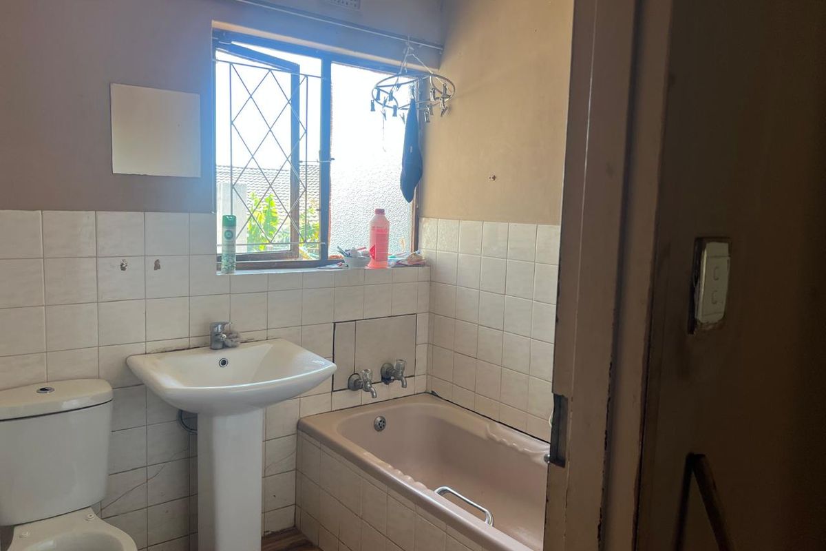 A functional bathroom featuring a pedestal basin, toilet, and built-in bathtub with tiled splashbacks and security burglar bars on the window.