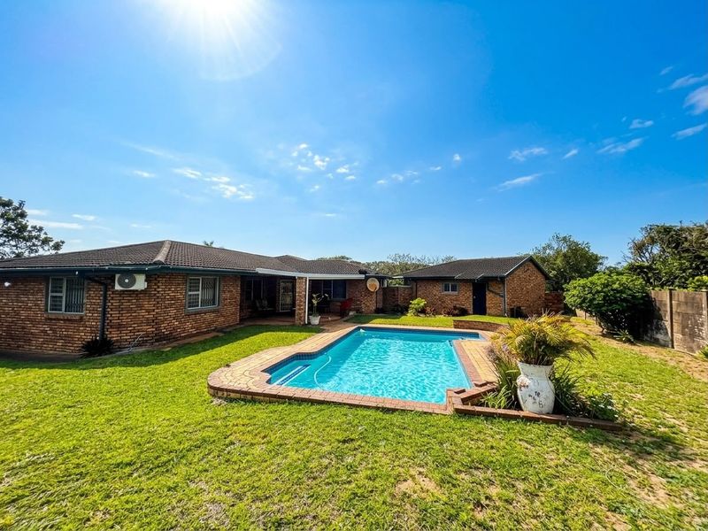 A spacious backyard featuring a swimming pool with brick coping, surrounded by a lawn and brick-built residential structures under a clear blue sky.