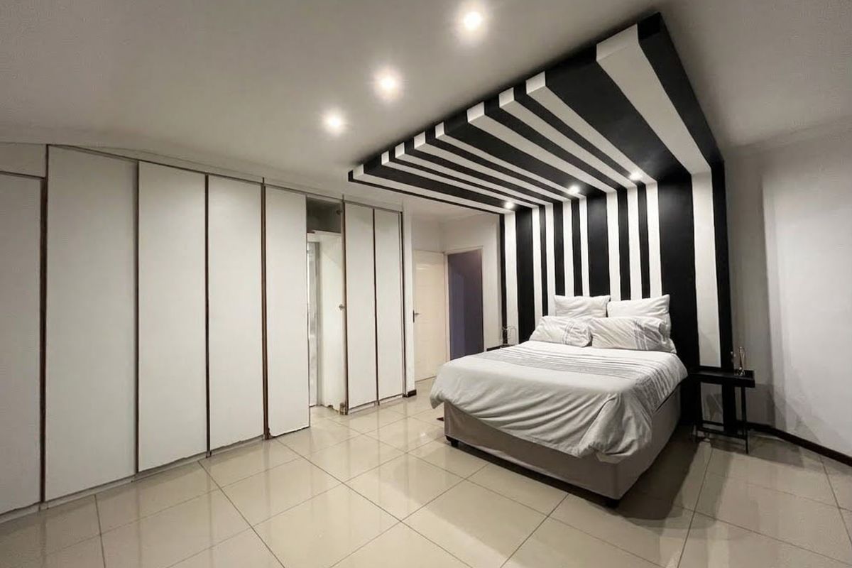 Spacious bedroom featuring large built-in cupboards, polished tile flooring, and a striking black and white decorative ceiling and wall feature.