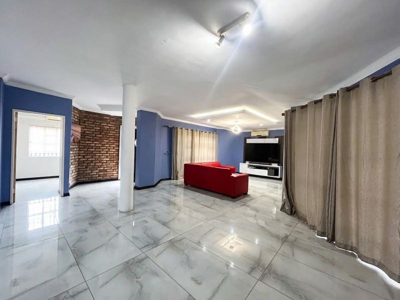 Spacious living room featuring polished marble-look porcelain tiles, a striking exposed brick feature wall, and a modern recessed tray ceiling with integrated lighting.