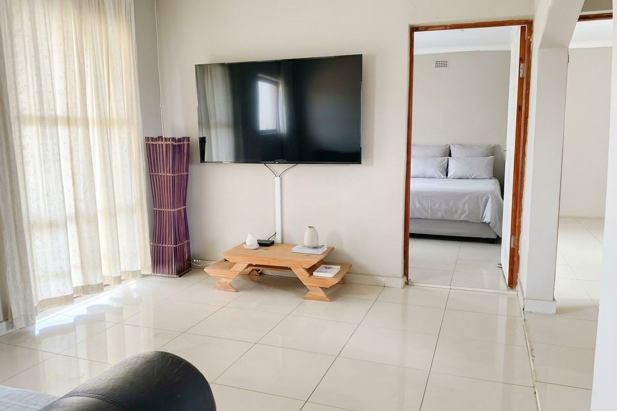 A bright living area featuring neutral tiled flooring and a view into an adjacent bedroom through a doorway.