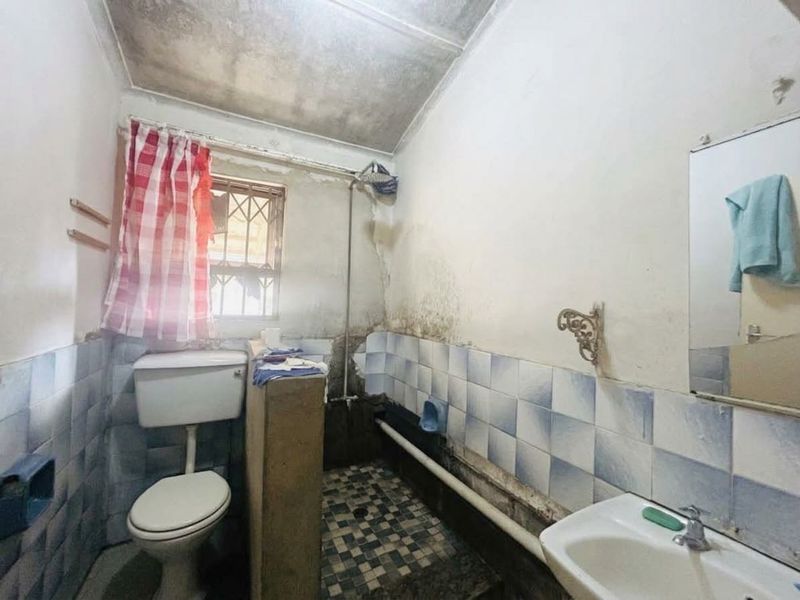 A dated bathroom requiring significant renovation, featuring a basic toilet, wall-mounted basin, and shower area with visible wall damage and surface deterioration.