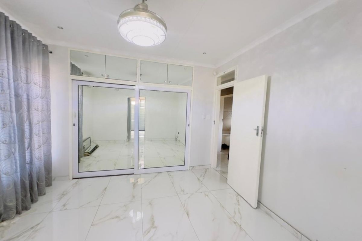 A bright, empty bedroom featuring modern white marble-look porcelain floor tiles, built-in cupboards with mirrored sliding doors, and a central ceiling light fixture.