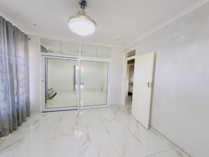 A bright, empty bedroom featuring modern white marble-look porcelain floor tiles, built-in cupboards with mirrored sliding doors, and a central ceiling light fixture.