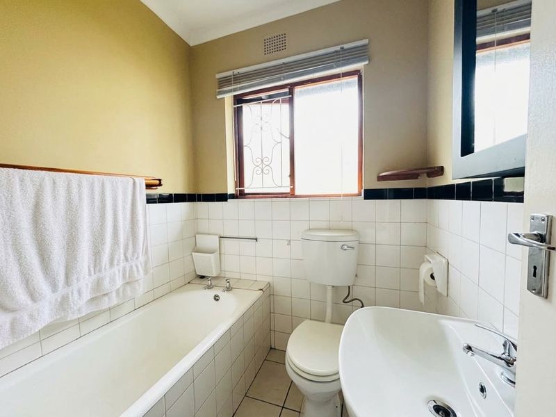 A functional bathroom featuring a bathtub, pedestal sink, and toilet with tiled finishes and a window with security bars.