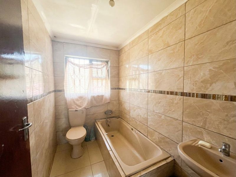 A compact bathroom featuring a bathtub, toilet, and washbasin with full-height wall tiling and security bars on the window.