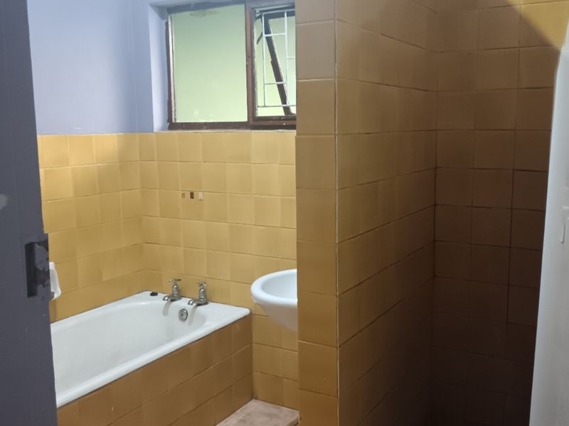 A dated bathroom featuring a built-in bathtub, wall-mounted basin, and yellow ceramic wall tiling. The space is vacant and ready for renovation.