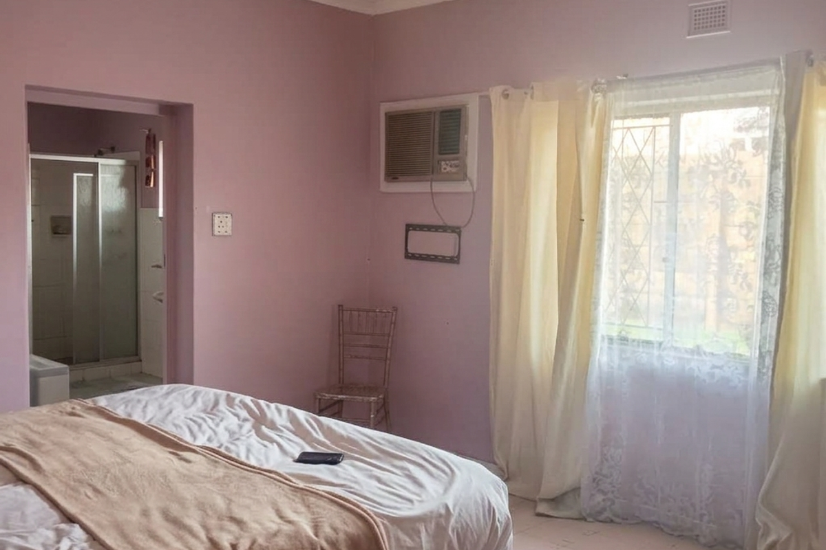 A bedroom featuring a ceiling fan, window-mounted air conditioner, and an en-suite bathroom. The room includes tiled flooring and a window with security bars.