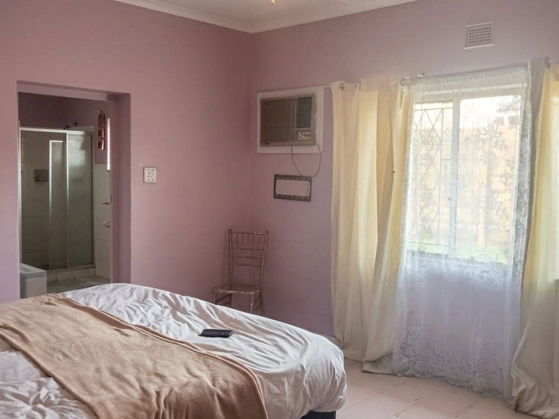 A bedroom featuring a ceiling fan, window-mounted air conditioner, and an en-suite bathroom. The room includes tiled flooring and a window with security bars.