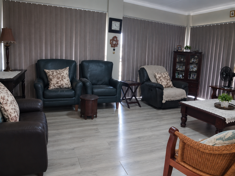 A spacious living room featuring modern light-toned laminate flooring, ceiling fan, and full-length vertical blinds covering the windows.