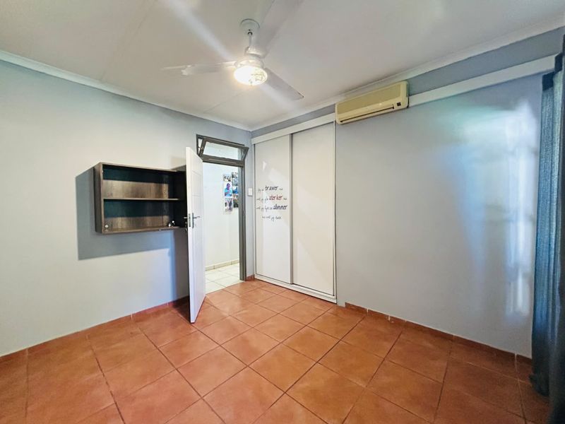A clean, empty bedroom featuring tiled flooring, a built-in sliding wardrobe, a ceiling fan, and a wall-mounted air conditioning unit.