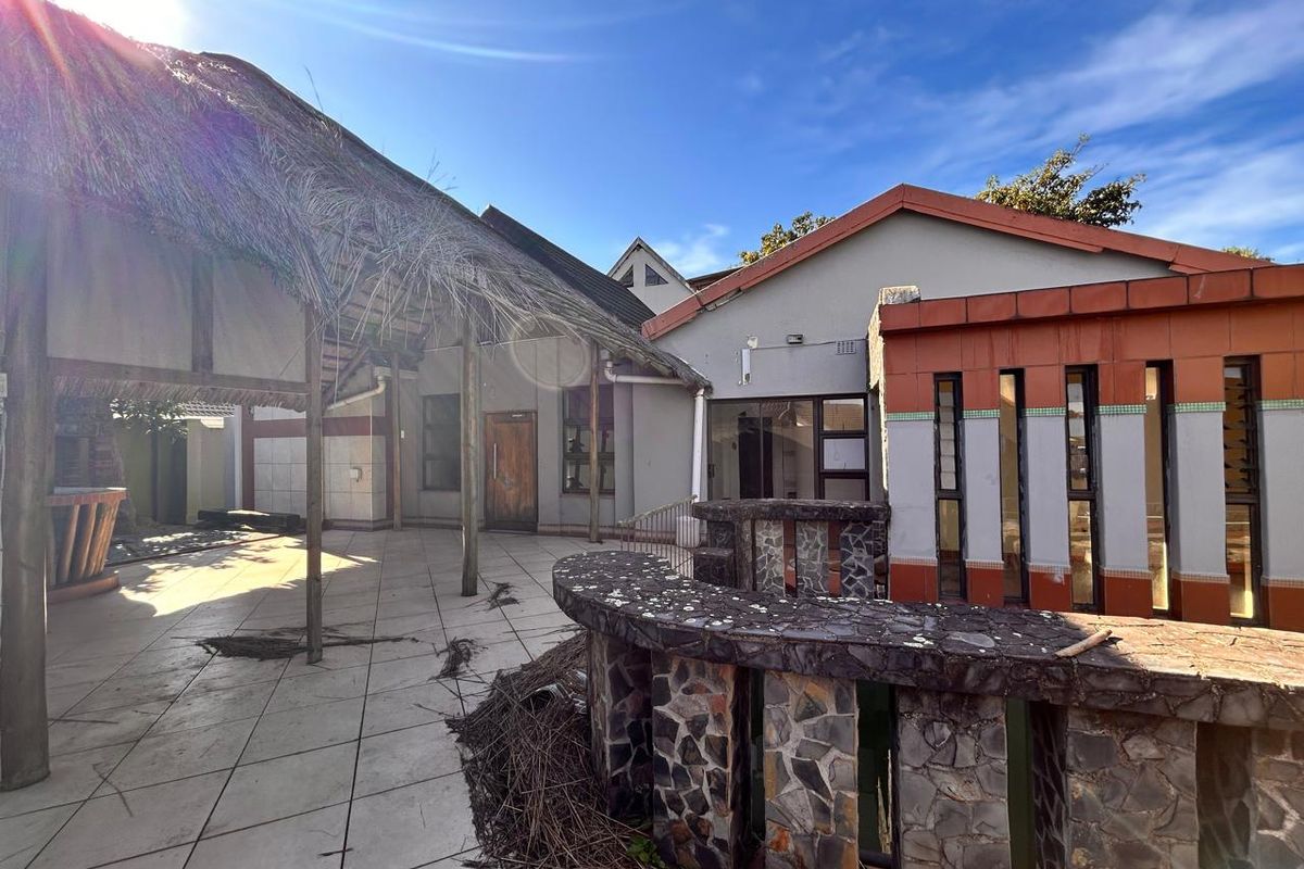 A rear exterior view featuring a covered patio area with a thatched roof, stone masonry accents, and tiled flooring leading to the main building structure.