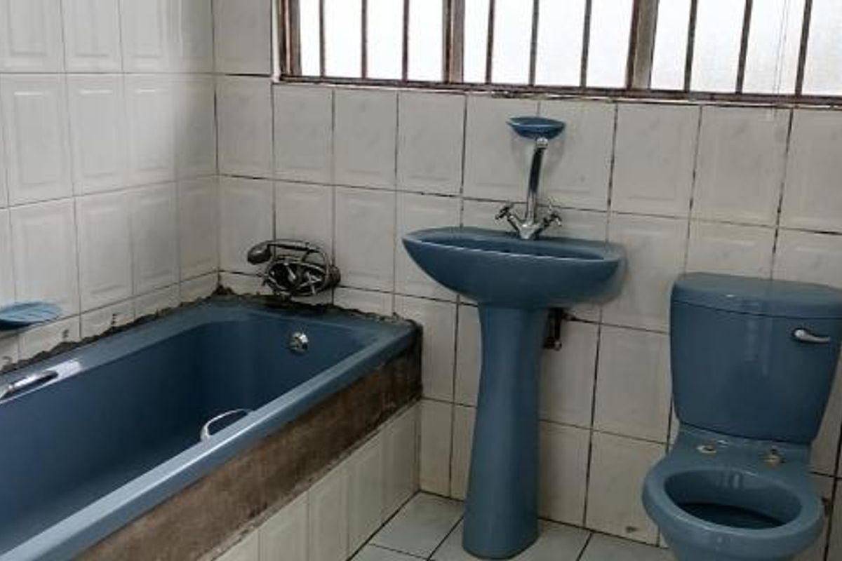 A dated bathroom featuring blue sanitaryware, including a bathtub, pedestal basin, and toilet, with white wall tiling and a security-barred window.