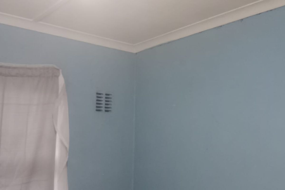 A simple bedroom interior featuring blue painted walls, ceiling coving, and a wall-mounted ventilation vent.
