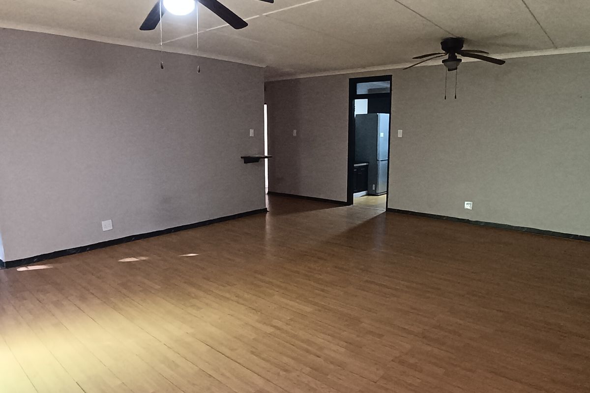 Spacious, empty living area featuring laminate flooring, ceiling fans, and a tiled ceiling, with a view into an adjacent room.