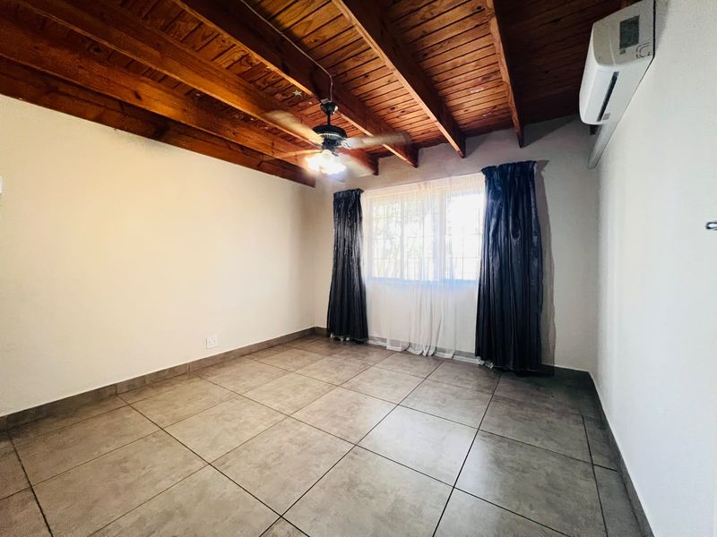A spacious bedroom featuring exposed timber beam ceilings, tiled flooring, a ceiling fan, and a wall-mounted air conditioning unit.