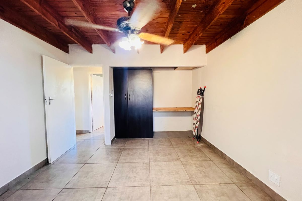 A bedroom featuring tiled flooring, an exposed timber beam ceiling with a ceiling fan, and a built-in wardrobe with an integrated desk area.