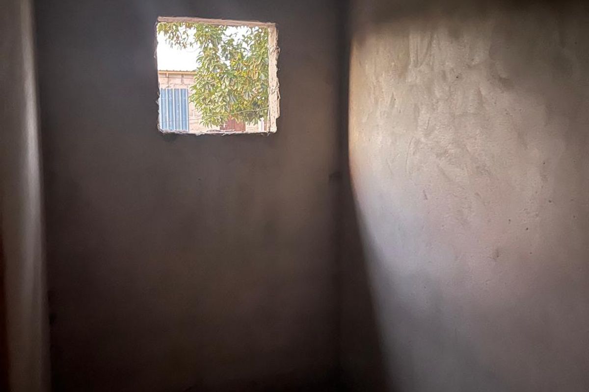 An unfinished room in shell condition featuring exposed rafters, cement rendered walls, and an open window aperture.