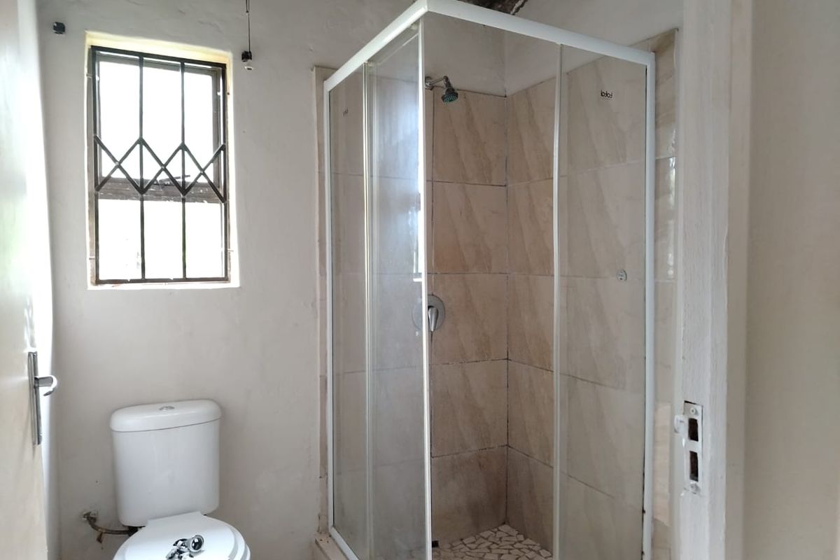 A functional bathroom featuring a glass-enclosed shower with tiled walls, a standard toilet, and a window with security bars.