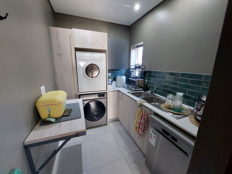 A functional laundry room featuring modern light wood cabinetry, a tiled backsplash, stainless steel sink, and space for integrated appliances.