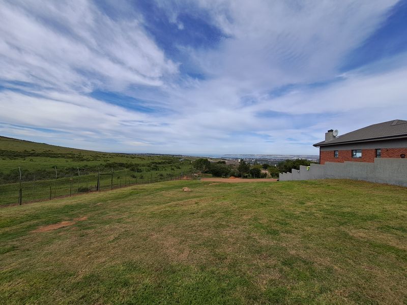 A wide-angle view of a vacant residential plot featuring a gentle slope, perimeter fencing, and a distant coastal landscape.