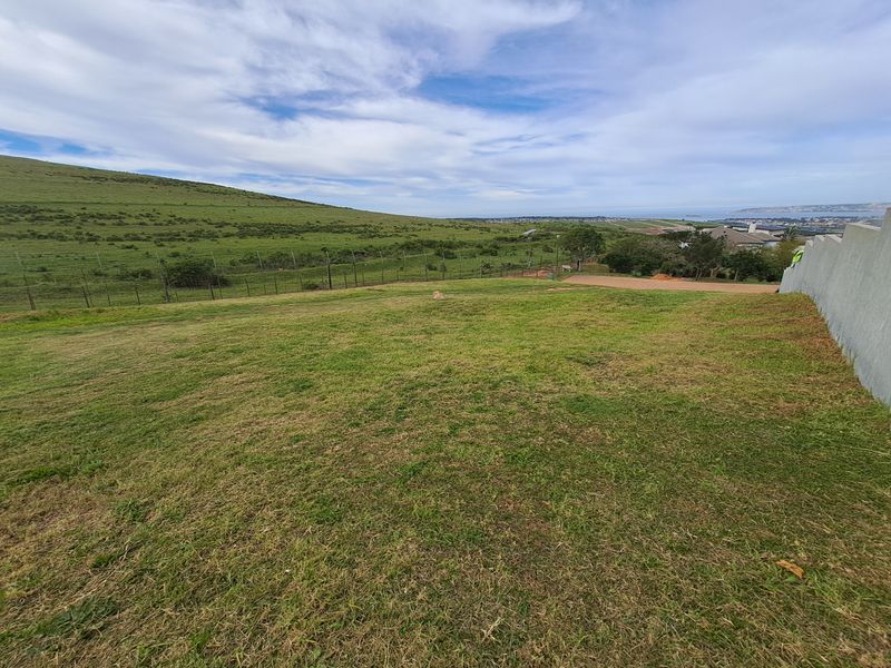 A wide-angle view of a vacant residential plot featuring grassy terrain, perimeter fencing, and a retaining wall with distant coastal views.