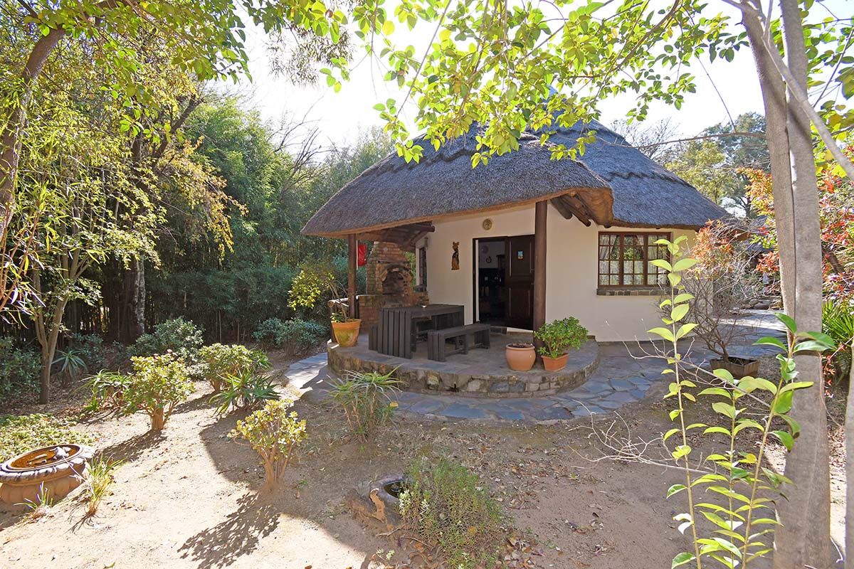 A charming thatched-roof lodge unit featuring a stone-paved patio with a built-in brick braai, nestled in a lush, mature garden setting.