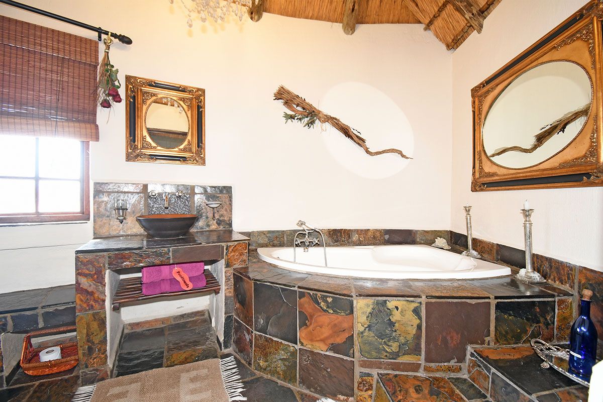 A rustic bathroom featuring a built-in corner bathtub and a stone-tiled vanity with a vessel sink, set under a traditional thatched roof.