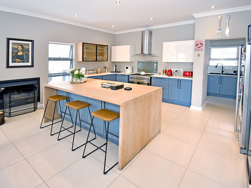 Modern open-plan kitchen featuring a large wooden island, blue cabinetry, stainless steel appliances, and an integrated fireplace, with an adjacent scullery area.