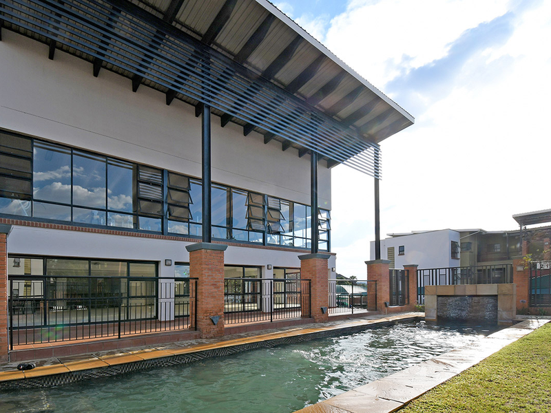 A well-maintained communal pool area featuring a water feature, brick pillars, and a modern metal roof structure overlooking the apartment complex.