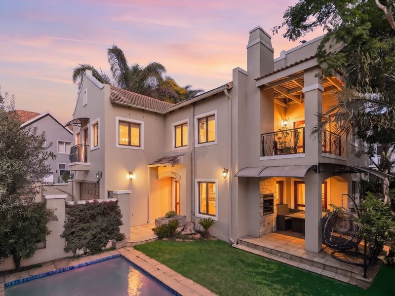 A beautifully presented double-story rear elevation featuring a swimming pool, covered patio with built-in braai, and an upper-level balcony at twilight.