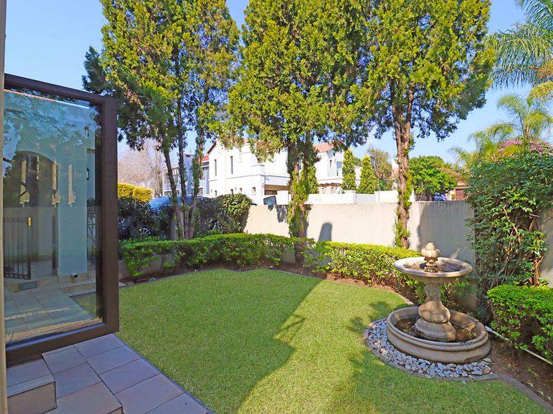 A well-maintained private garden featuring a manicured lawn, mature trees, a stone water fountain, and a tiled patio area.