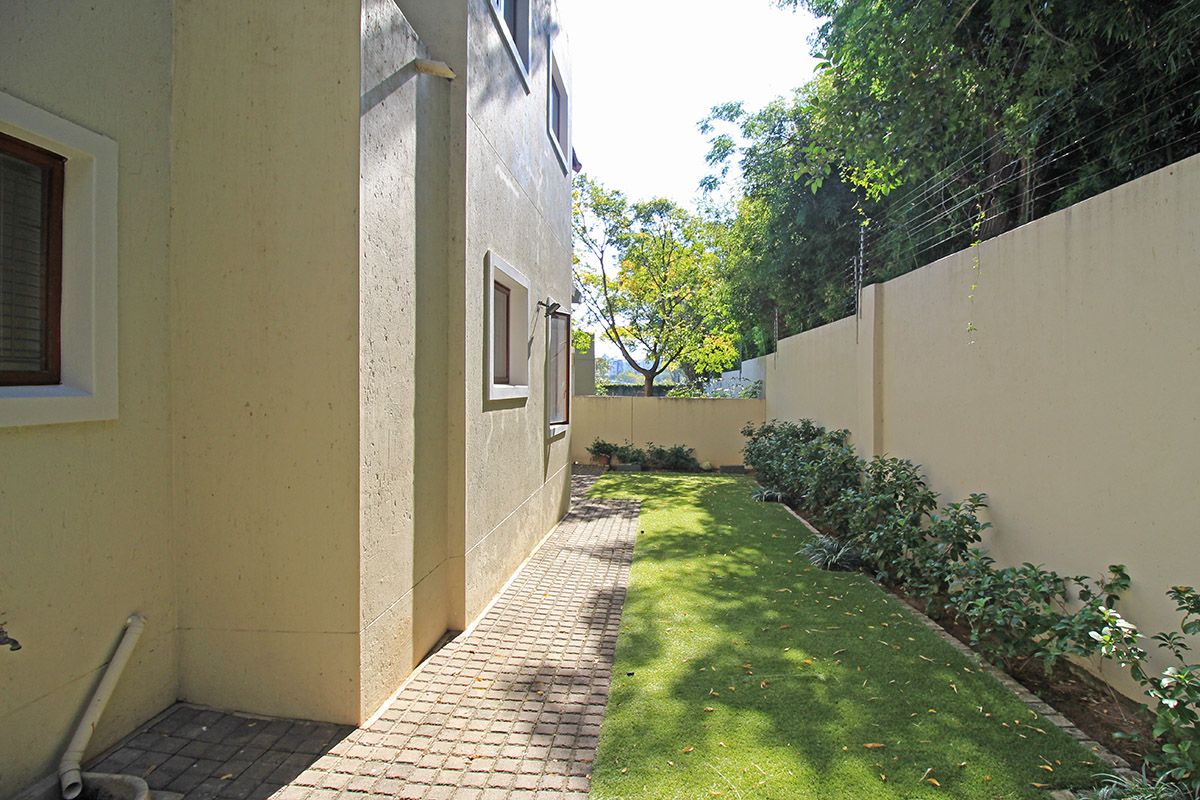 A side garden area featuring a paved walkway, well-maintained lawn, and high boundary walls with integrated electric fencing for added security.