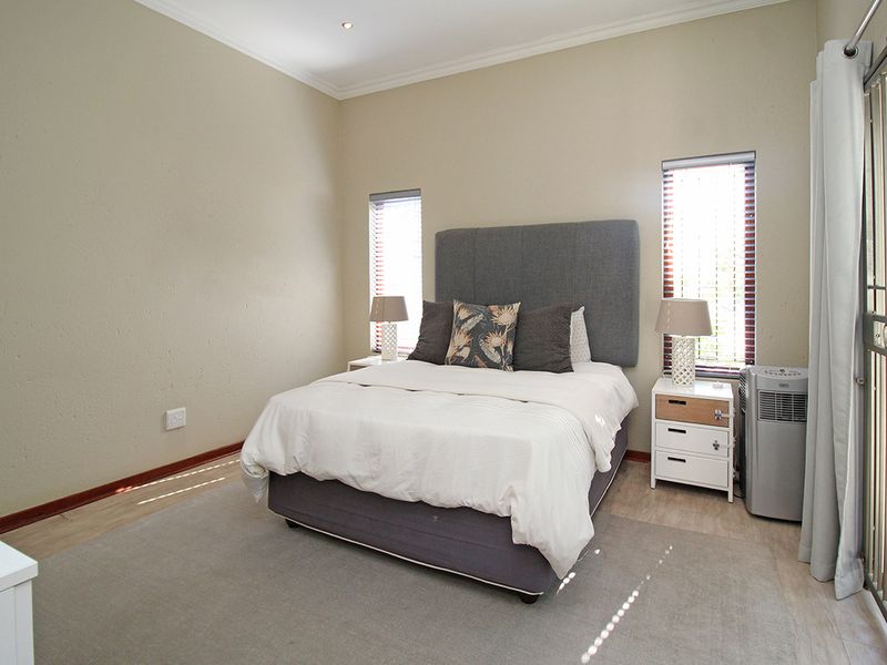 A well-maintained bedroom featuring laminate flooring, recessed downlights, and dual windows with venetian blinds, providing access to an outdoor area via a security gate.