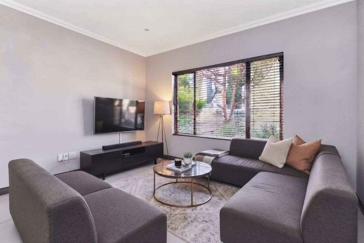 A well-maintained living room featuring neutral wall tones, tiled flooring, recessed lighting, and large windows fitted with venetian blinds.