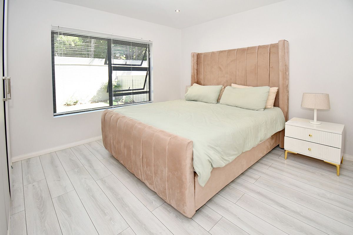 A bright and modern bedroom featuring light-toned wood-look flooring, recessed ceiling lighting, and a large window with integrated blinds.