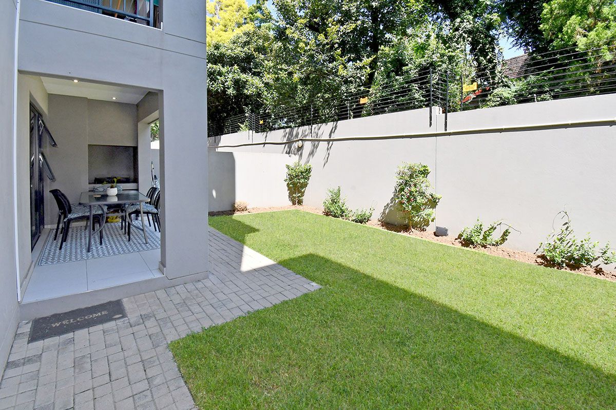 A private rear garden featuring a paved patio area with a built-in braai, adjacent to a well-maintained lawn and perimeter wall.