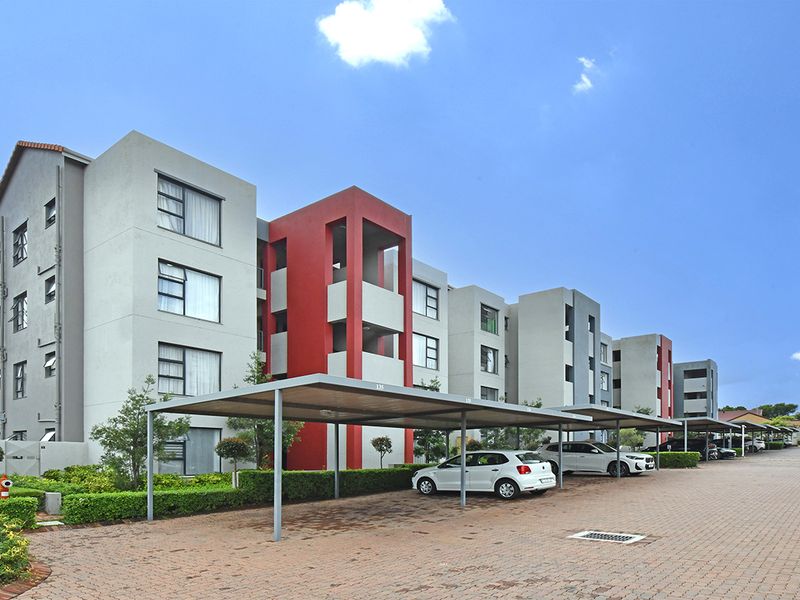 Exterior view of a modern multi-story apartment complex featuring covered parking bays, paved driveway, and well-maintained landscaped hedging.