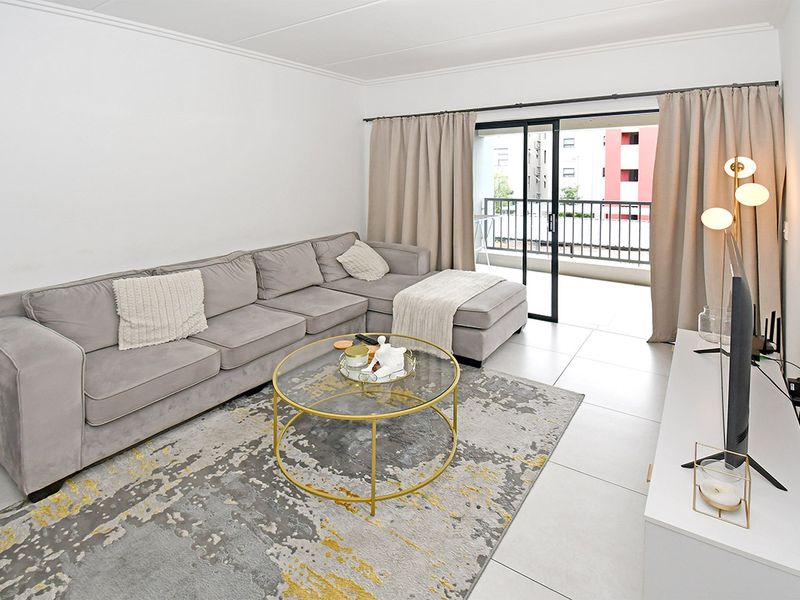 A bright and well-maintained living room featuring neutral tiled flooring, cove cornicing, and direct access to a private balcony through sliding glass doors.