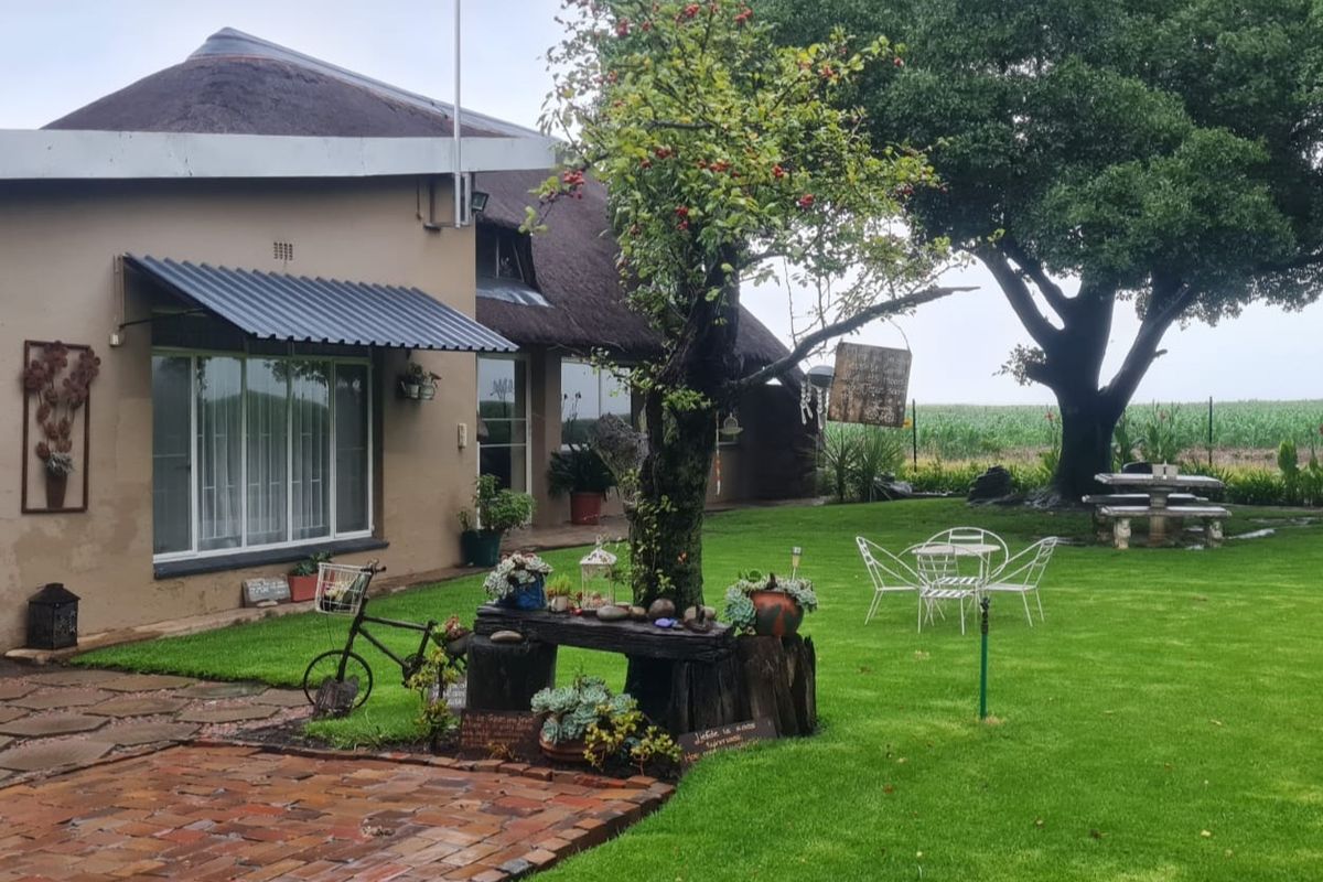 A well-maintained rear garden area featuring a thatched roof dwelling, brick paved patio, and expansive lawn with mature trees.