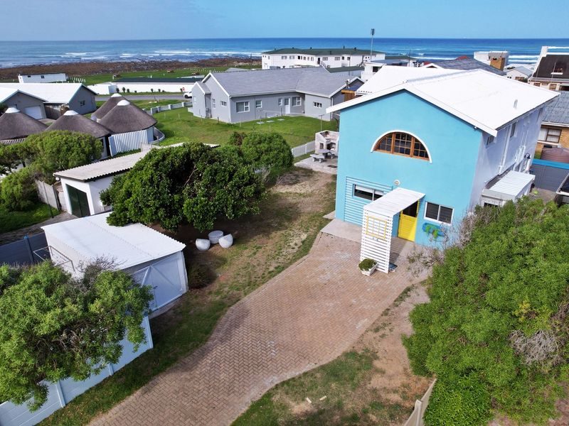 Aerial view of a coastal property featuring a blue exterior, paved driveway, detached garage, and built-in outdoor braai area near the ocean.