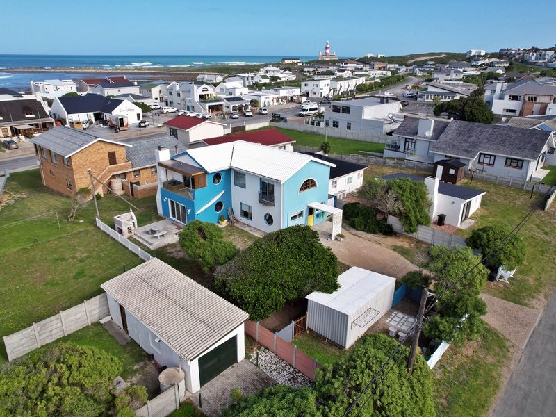 Aerial view of a coastal property featuring a blue two-story house, detached garage, paved driveway, and outdoor entertainment area near the ocean.