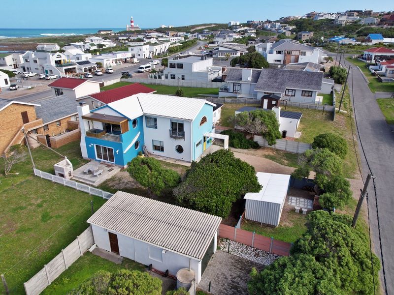 Aerial view of a coastal residential property featuring a blue two-story house, detached garage, paved driveway, and garden area in a suburban setting.