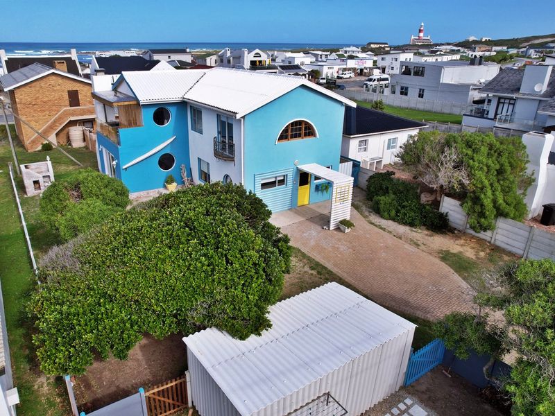 Aerial view of a coastal property featuring a blue exterior, corrugated metal roof, paved driveway, and balcony, situated in a residential seaside neighborhood.
