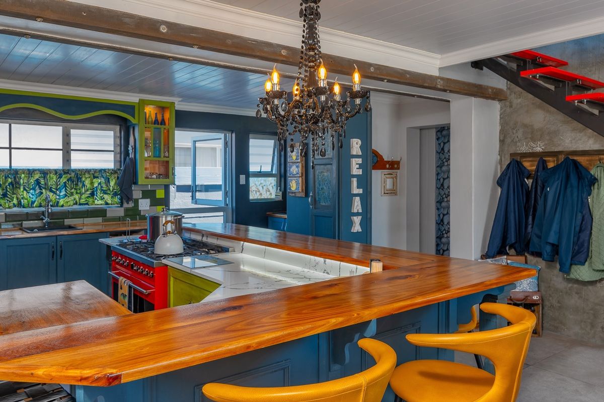 A vibrant kitchen featuring custom blue and green cabinetry, a large timber breakfast bar, stone countertops, and a striking central chandelier.