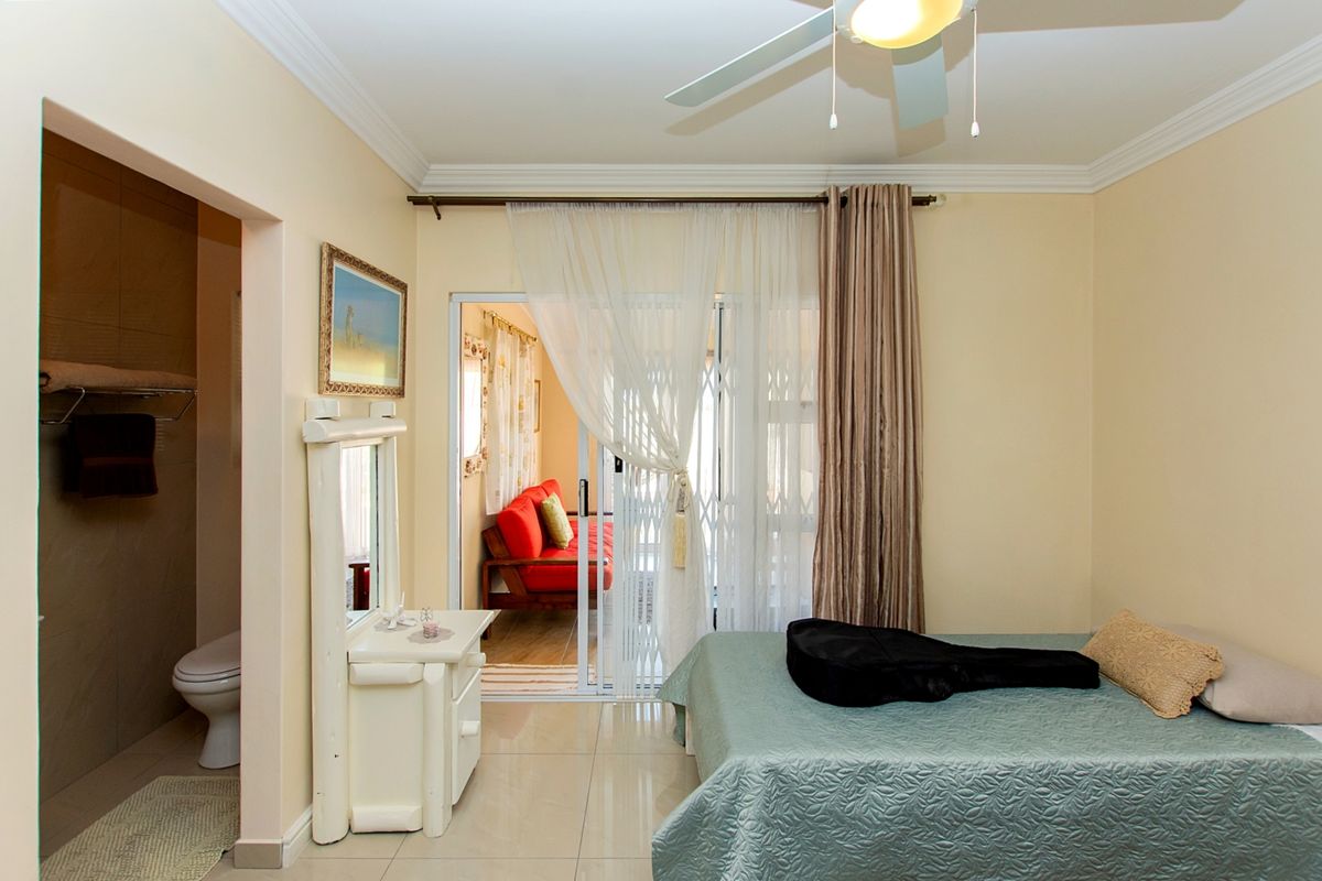 A well-lit bedroom featuring tiled flooring, crown molding, a ceiling fan, and direct access to an en-suite bathroom and an adjoining living space.